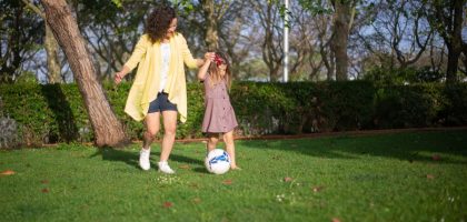 A mother and daughter enjoying a sunny day playing soccer in a lush green park.