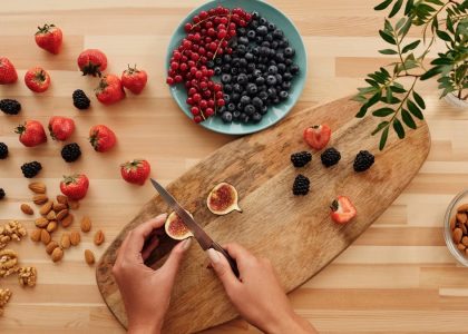 Overhead shot of fresh fruits, nuts, and hands preparing ingredients on a wooden board.
